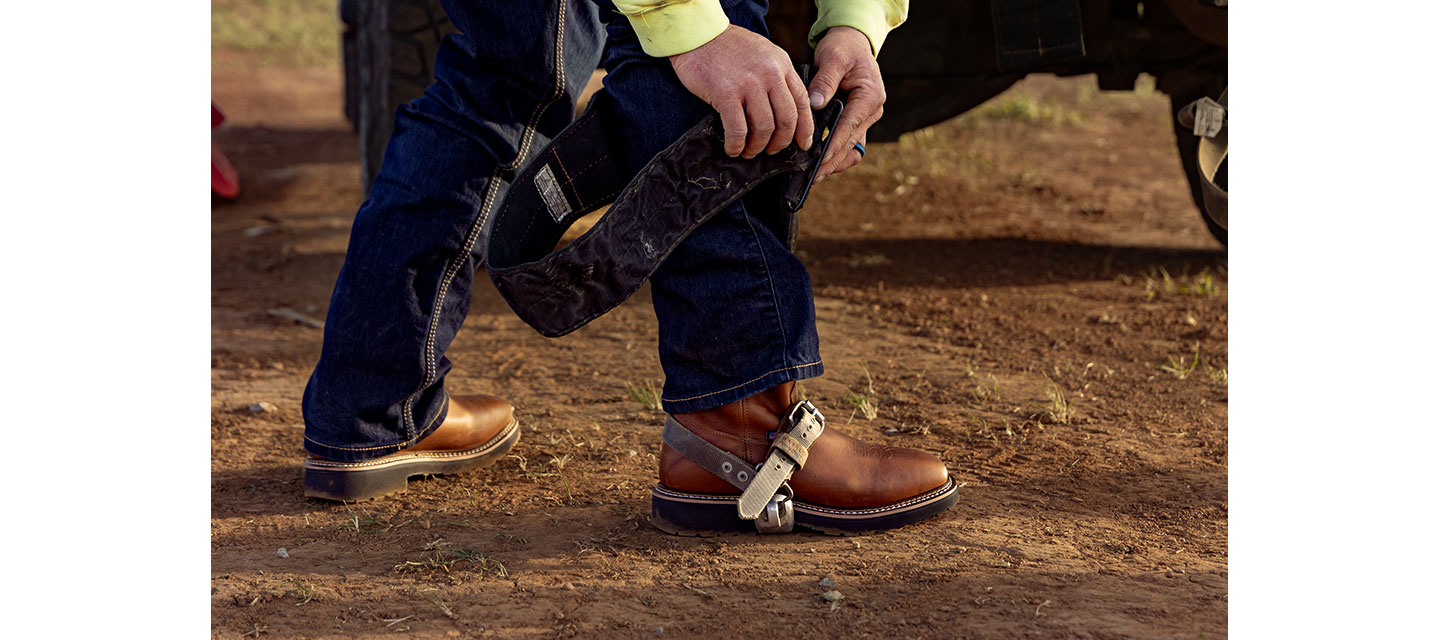 Close-up of a lineman, Dylan Edgmon, wearing brown leather work boots and attaching climbing gear while standing on dirt.